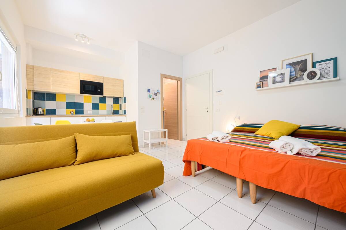 Une chambre d’hôtel moderne avec un canapé jaune, un lit orange, un coin cuisine et une étagère murale où sont exposées des photos.