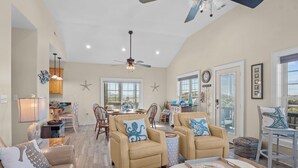 A spacious living room with beige sofas, a dining area with wooden chairs, and a ceiling fan. - Soundside Home with Elevator, Watersports Gear, Community Marina, Beach Nearby (Hatteras)