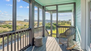 A covered porch with two wicker chairs and a small round table, overlooking a grassy area with houses in the distance. - Soundside Home with Elevator, Watersports Gear, Community Marina, Beach Nearby (Hatteras)