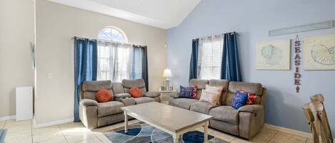 Ein Wohnzimmer mit zwei Sofas, einem Couchtisch und einem Deckenventilator. Vor den Fenstern hängen Vorhänge, und an der Wand ist ein Schild mit der Aufschrift 'SEASIDE!'.