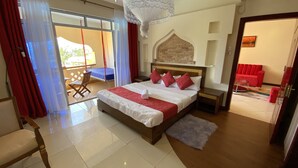 A hotel room with a bed, a wooden nightstand, a red sofa, a balcony with a chair, and a view of the outside. - Lux Suites Eden Seafront Apartments (Mombasa)