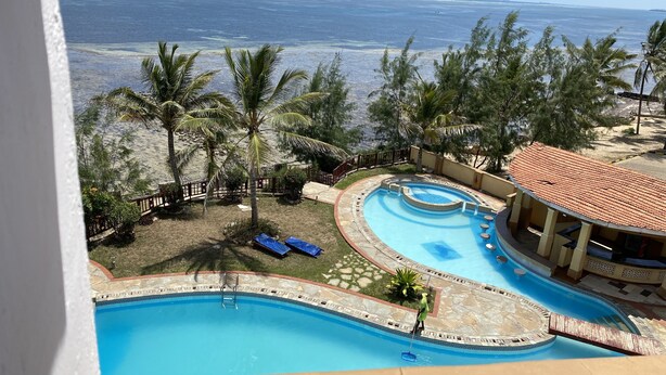 A swimming pool with a curved design, surrounded by palm trees and a view of the ocean. - Lux Suites Eden Seafront Apartments (Mombasa)