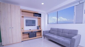 Ein modernes Wohnzimmer mit einem grauen Sofa, einem Fernseher auf einer Holz-Unterhaltungsanlage und einem großen Fenster mit Blick auf die Stadt.