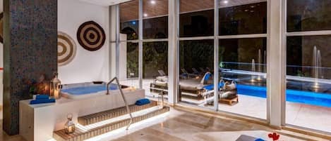 A modern indoor pool area with a hot tub, lounge chairs, and a view of an outdoor pool at night.