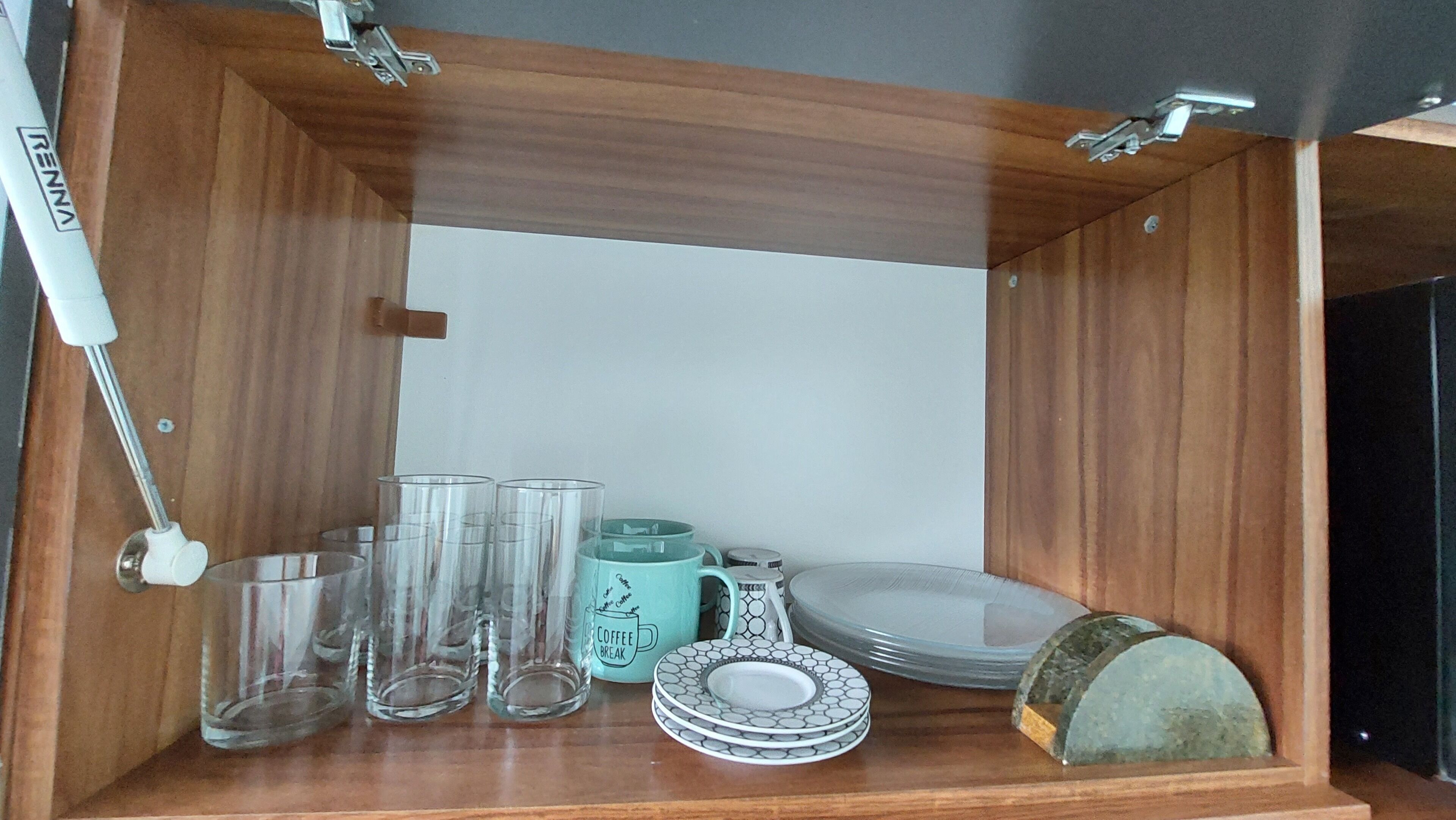 A wooden cabinet with a white interior, containing clear drinking glasses, a turquoise mug, a white mug, and a stack of plates.