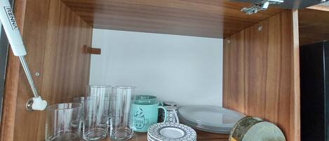 A wooden cabinet with a white interior, containing clear drinking glasses, a turquoise mug, a white mug, and a stack of plates.