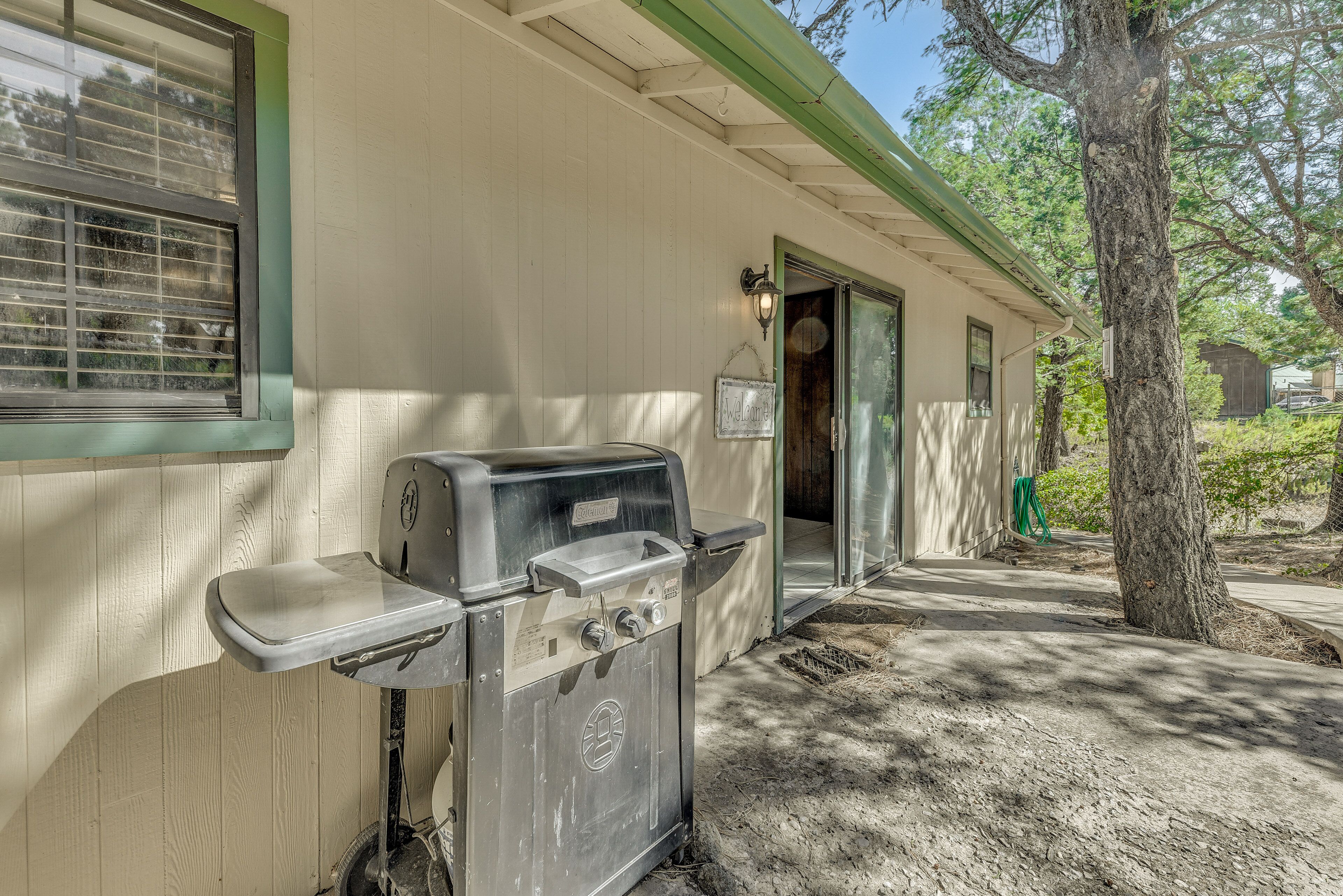 A portable grill attached to a house's exterior wall, a window with blinds, and a tree in the background.