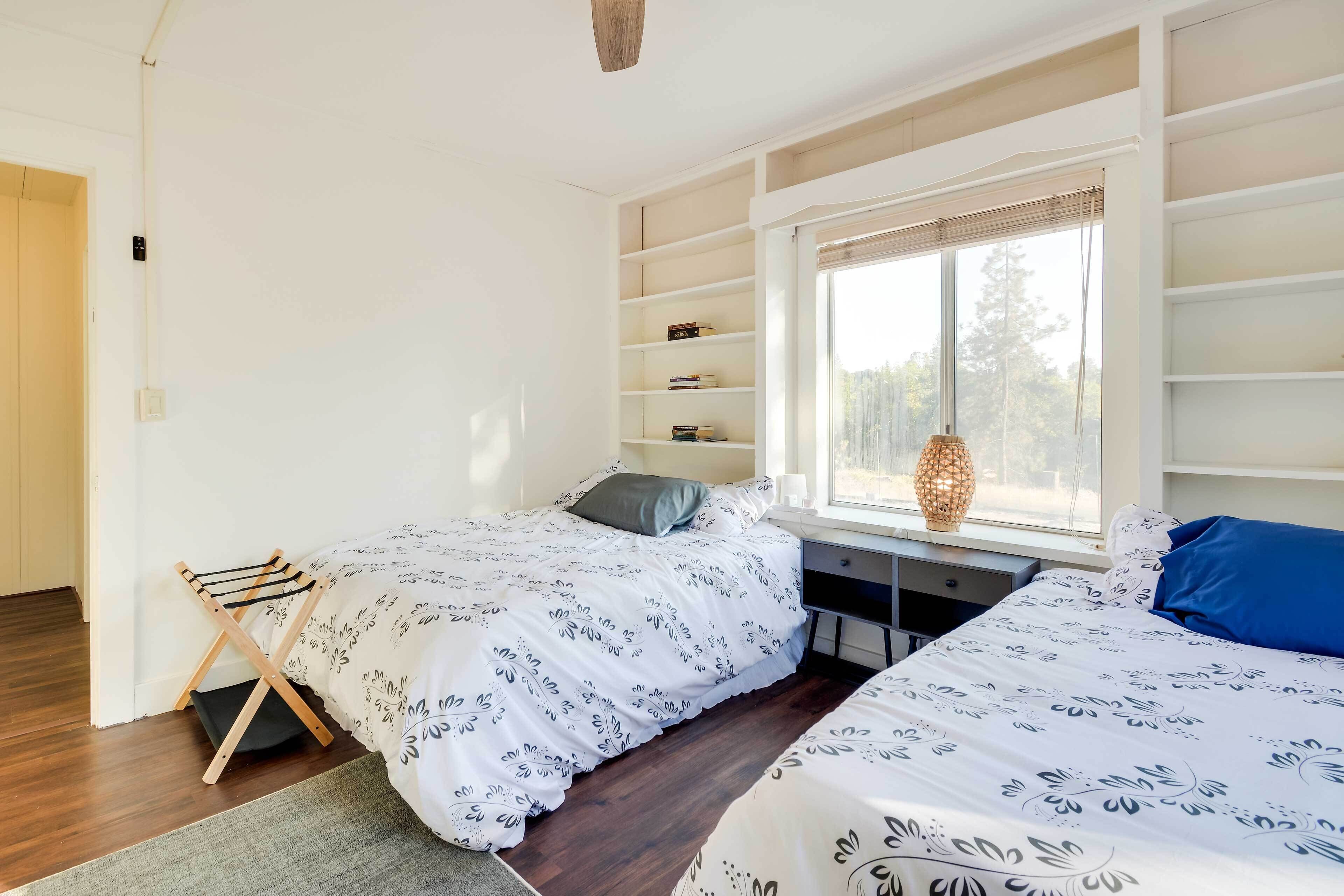 A bedroom with two beds, a window with blinds, a bedside table, and built-in shelving.