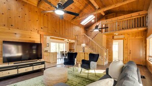 A spacious living room with a large flat-screen TV, comfortable seating, a staircase, and wooden walls. - Waterfront Mariposa Vacation Rental w/ Hot Tub (Mariposa)