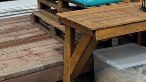 Un banco de madera con cojines turquesa y un recipiente de plástico sobre una terraza de madera.