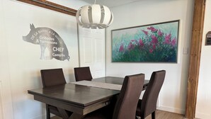 Un comedor con una mesa y sillas de madera oscura, una lámpara colgante blanca, un letrero de parrilla montado en la pared y un cuadro floral enmarcado.