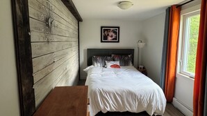 Un dormitorio con cabecera de madera, cama con ropa de cama blanca, mesita de noche, lámpara de noche y ventana con cortinas.