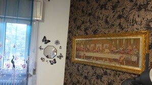 Une pièce dans laquelle on trouve un grand tableau représentant «La Cène», accroché au mur, une fenêtre ornée de rideaux à motifs floraux et un radiateur.