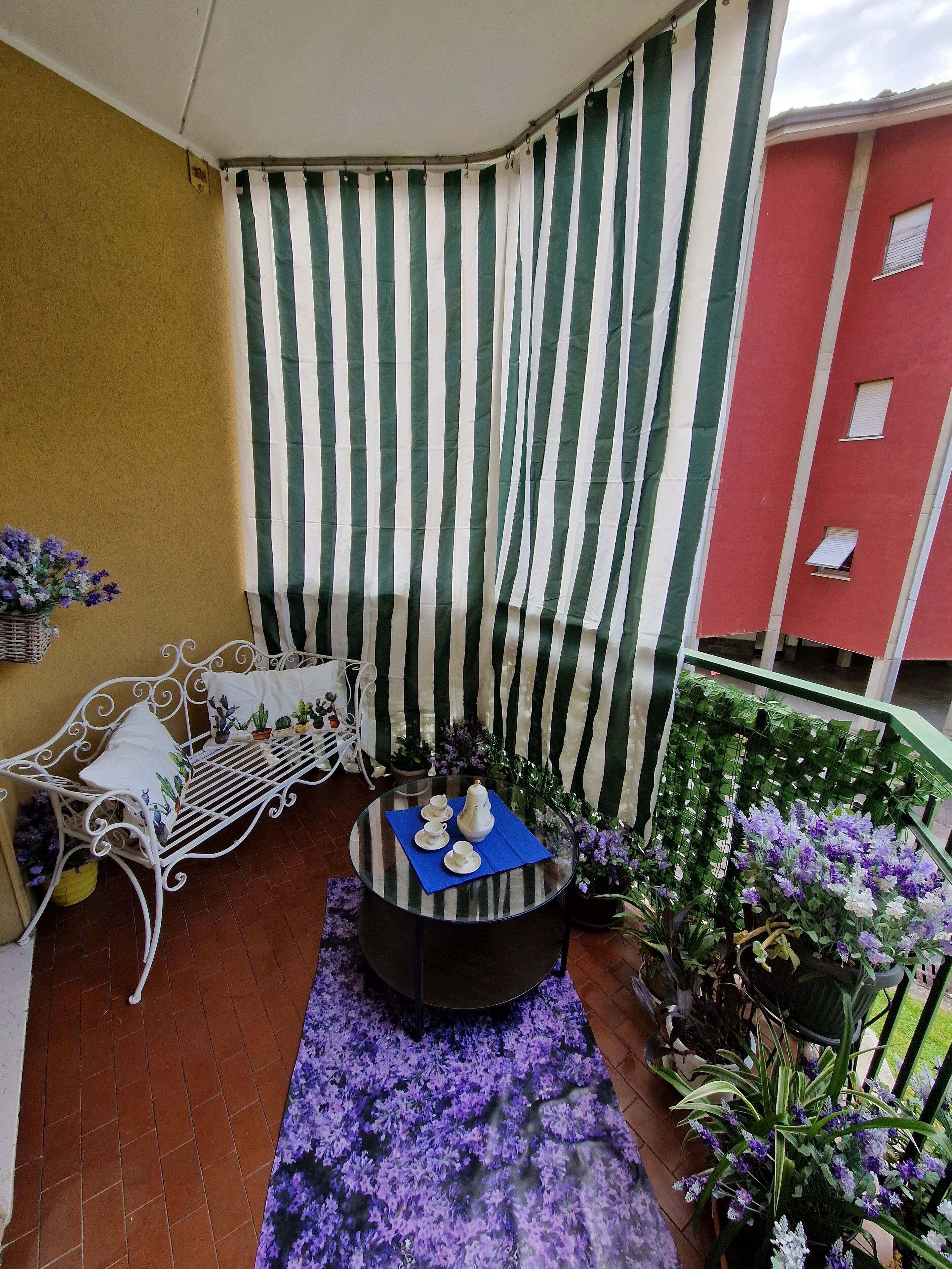 En lille balkon med en metalstol, et glasbord med tepotte og kopper, og et iøjnefaldende stribet gardin.