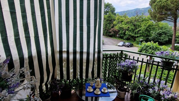 Un balcon avec un rideau à rayures, une table avec une théière et des tasses, des plantes en pot et une vue sur une route et la verdure.