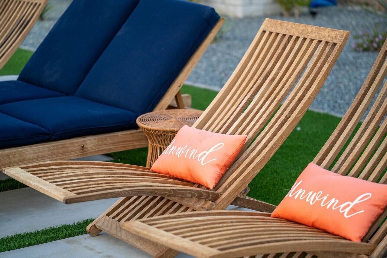 Two wooden lounge chairs with cushions, one blue and one orange, on a concrete surface with grass in the background.