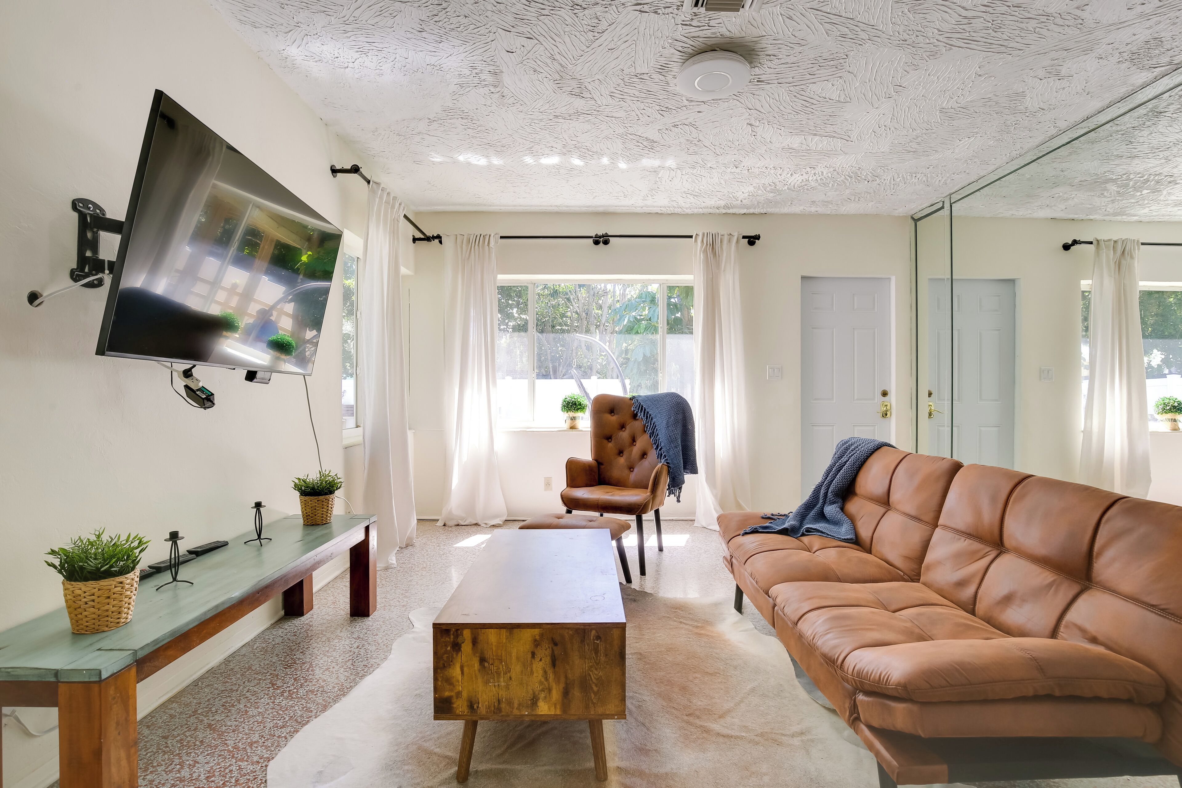 A living room with a brown leather sofa, a wooden coffee table, a TV mounted on the wall, and a bench with potted plants.