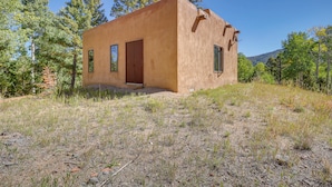 A small, single-story building with a flat roof, surrounded by a natural landscape with trees and a clear sky. - Pet-friendly Mountain Cabin in Taos County! (Amalia)