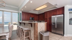 A modern kitchen with a marble island, wooden stools, a stainless steel refrigerator, and a view of the ocean.