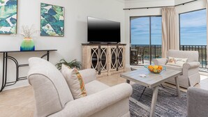A modern living room with a flat-screen TV, a dining table with a fruit bowl, and a balcony with a view. - Book Phoenix 7 Unit 7109 (Orange Beach)