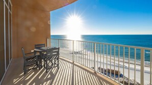 Een balkon met een tafel en stoelen, met uitzicht op een strand en een helder zicht op de oceaan, terwijl de zon schijnt.