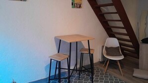 Une pièce avec une table en bois, deux tabourets, une chaise blanche et un escalier.
