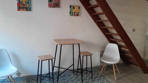 Une pièce avec un escalier en bois, une petite table accompagnée de deux tabourets et trois tableaux accrochés au mur.