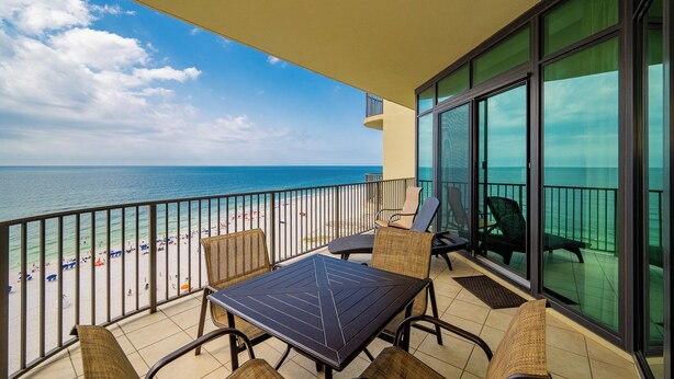 A balcony with a table and chairs, overlooking a beach with umbrellas and people. - Gulf Front Condo - Phoenix West 905 (Orange Beach)