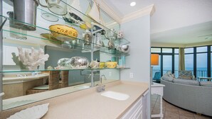 A modern bathroom with a glass shelf holding various decorative items, a large mirror, and a view of the ocean through large windows. - Gulf Front Condo - Phoenix West 905 (Orange Beach)