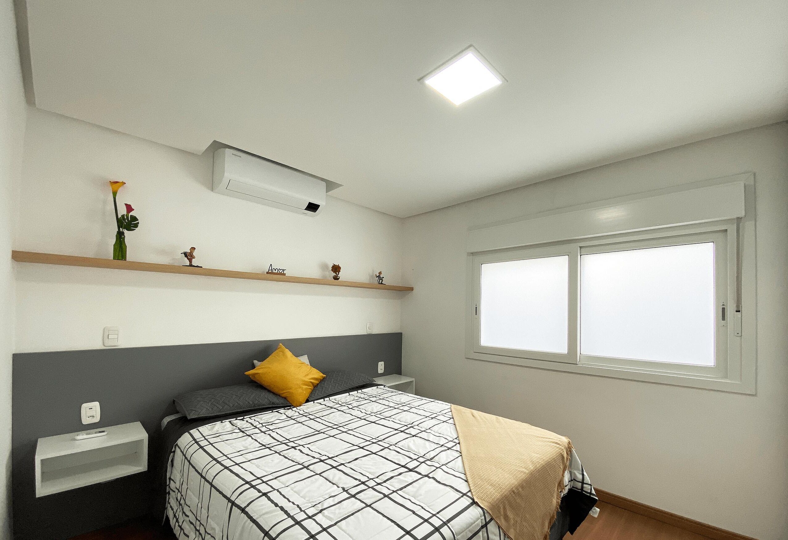 A bedroom with a bed, a nightstand, a shelf with decorative items, a window, and an air conditioning unit.
