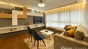 A modern living room with a sofa, chairs, a TV, and built-in shelves. - Apartamento Morada do Sol Gramado (Gramado)