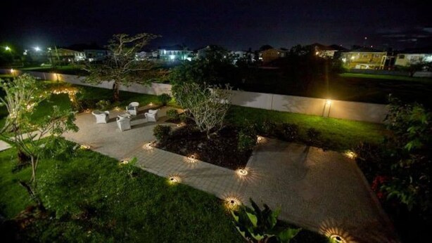 Een terras buiten met zitplaatsen en sfeerverlichting, omgeven door groen en een geplaveid pad.