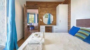 Een slaapkamer met een houten plafond, een groot bed met wit beddengoed, een nachtkastje met een spiegel, en zicht op een andere kamer door een geopende deur.