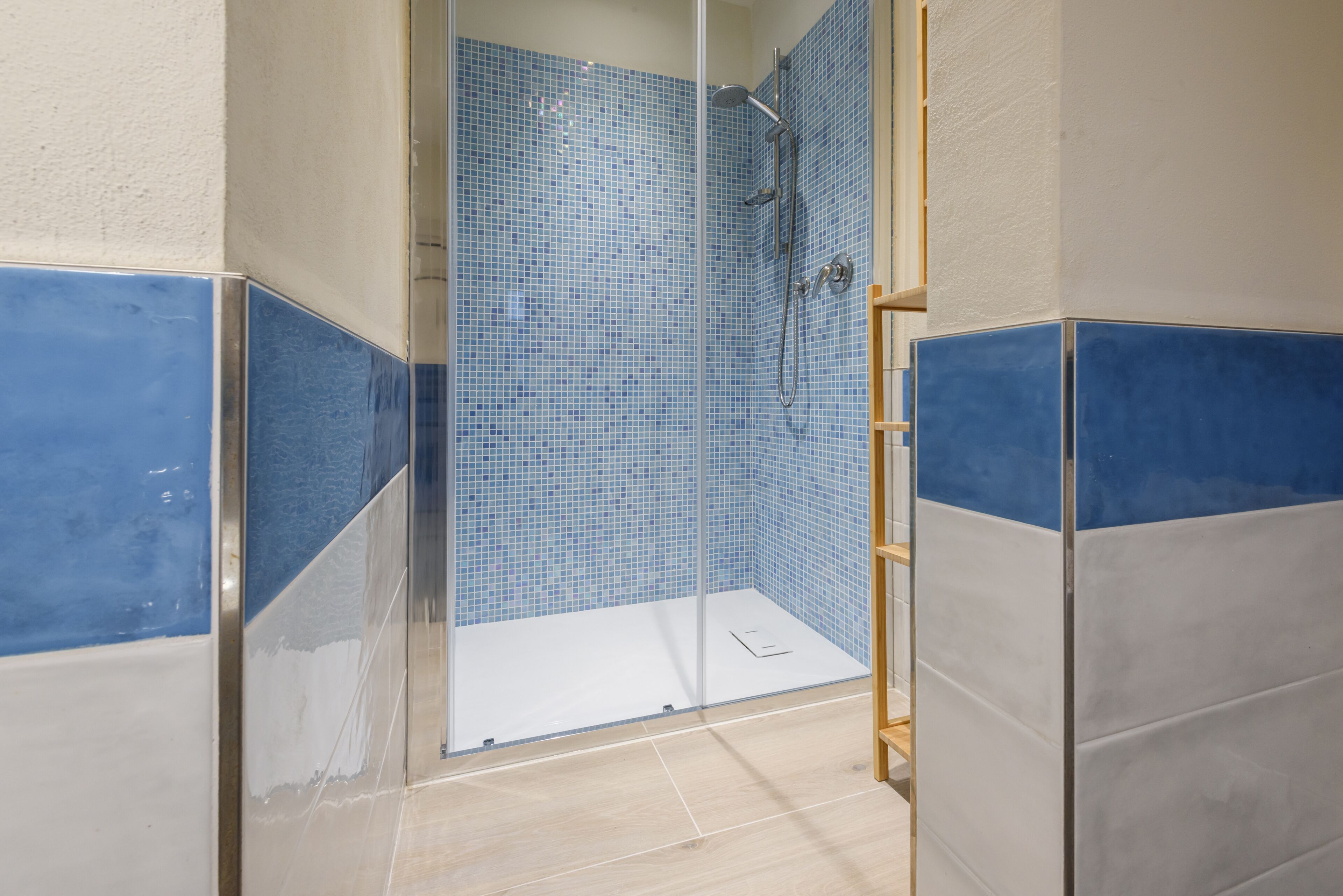 Zona de ducha con azulejos de mosaico azules, puerta de cristal y banco de madera.