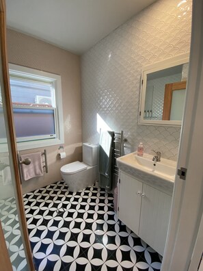 Une salle de bain avec des toilettes, un lavabo et un miroir. Il y a une fenêtre donnant sur l’extérieur. Le sol est recouvert d’un motif géométrique noir et blanc.