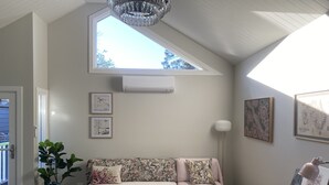 Un salon chaleureux avec un plafond haut, une lucarne, un canapé agrémenté de coussins à motifs floraux, deux petites tables d’appoint rondes en bois et un lampadaire.