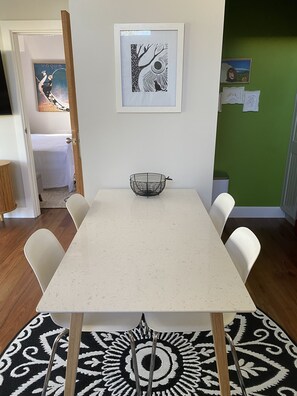 Un coin repas avec une table blanche, quatre chaises blanches, un tapis à motifs noir et blanc et une œuvre d’art encadrée accrochée au mur.