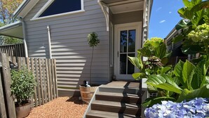 Une maison à l’extérieur grise, avec un porche à l’avant doté d’un escalier en bois, une petite plante en pot et une allée gravillonnée.