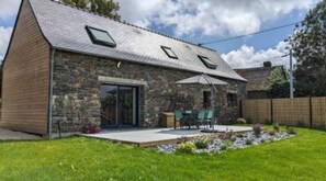 Un edificio de piedra con techo inclinado, un patio con mobiliario exterior y un jardín con césped.