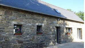 Un bâtiment en pierre avec un toit en pente, trois fenêtres ornées de jardinières et une allée gravillonnée.