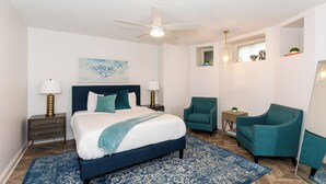 A modern bedroom with a large bed, a ceiling fan, a blue patterned rug, and teal armchairs. - Chic Condo Near Cedar Point (Sandusky)