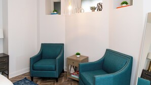 A bedroom with two teal armchairs, a wooden floor, a blue patterned rug, a small bedside table, a mirror, and a white bed. - Chic Condo Near Cedar Point (Sandusky)