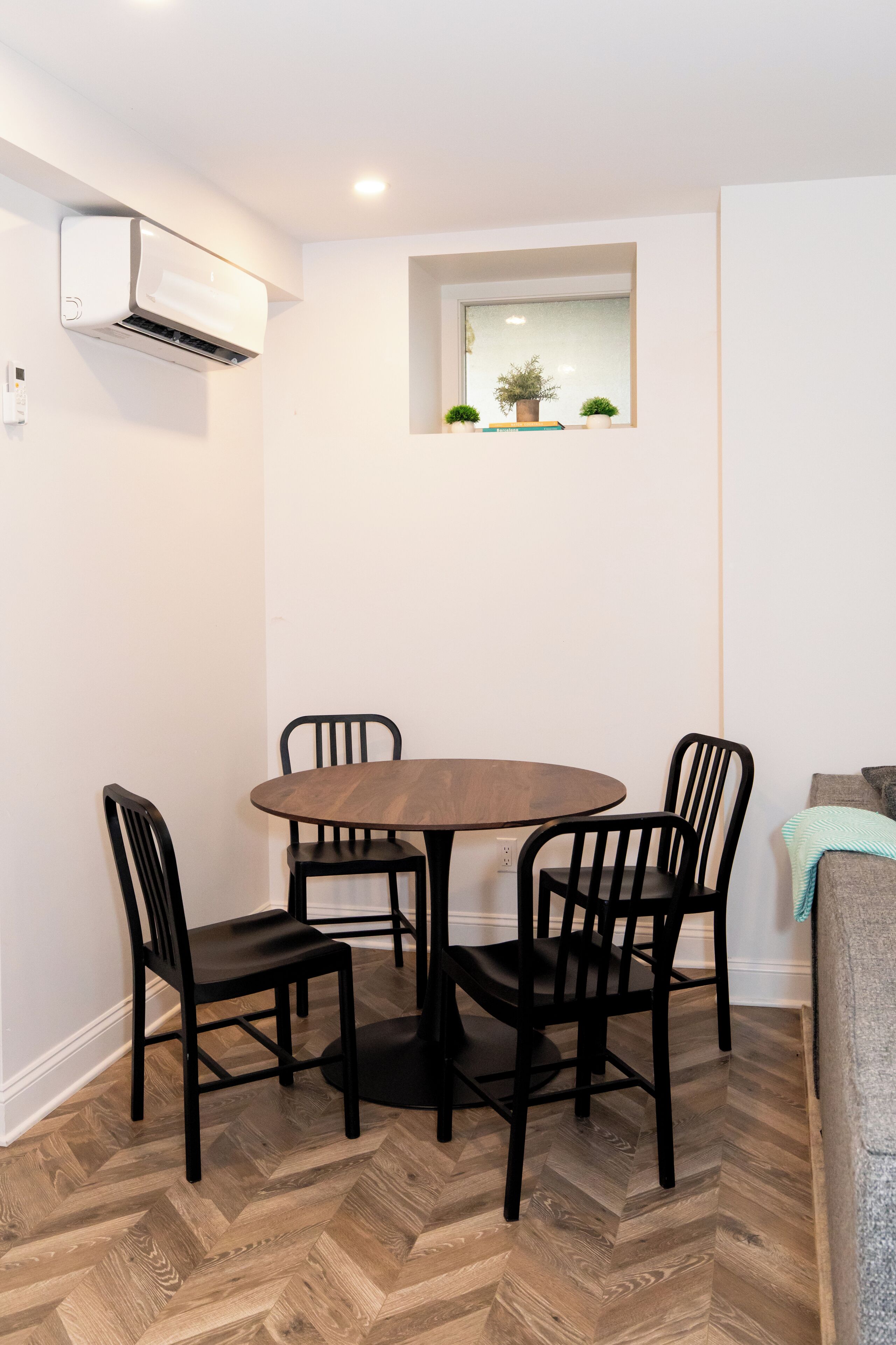 A dining area with a round table and four chairs, an air conditioning unit, a small window with plants, and a herringbone-patterned floor.