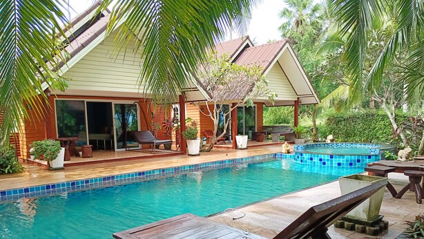 A pool area with a wooden deck, lounge chairs, and a house with a tiled roof and large windows. - Sunrise Resort (Bang Saphan)