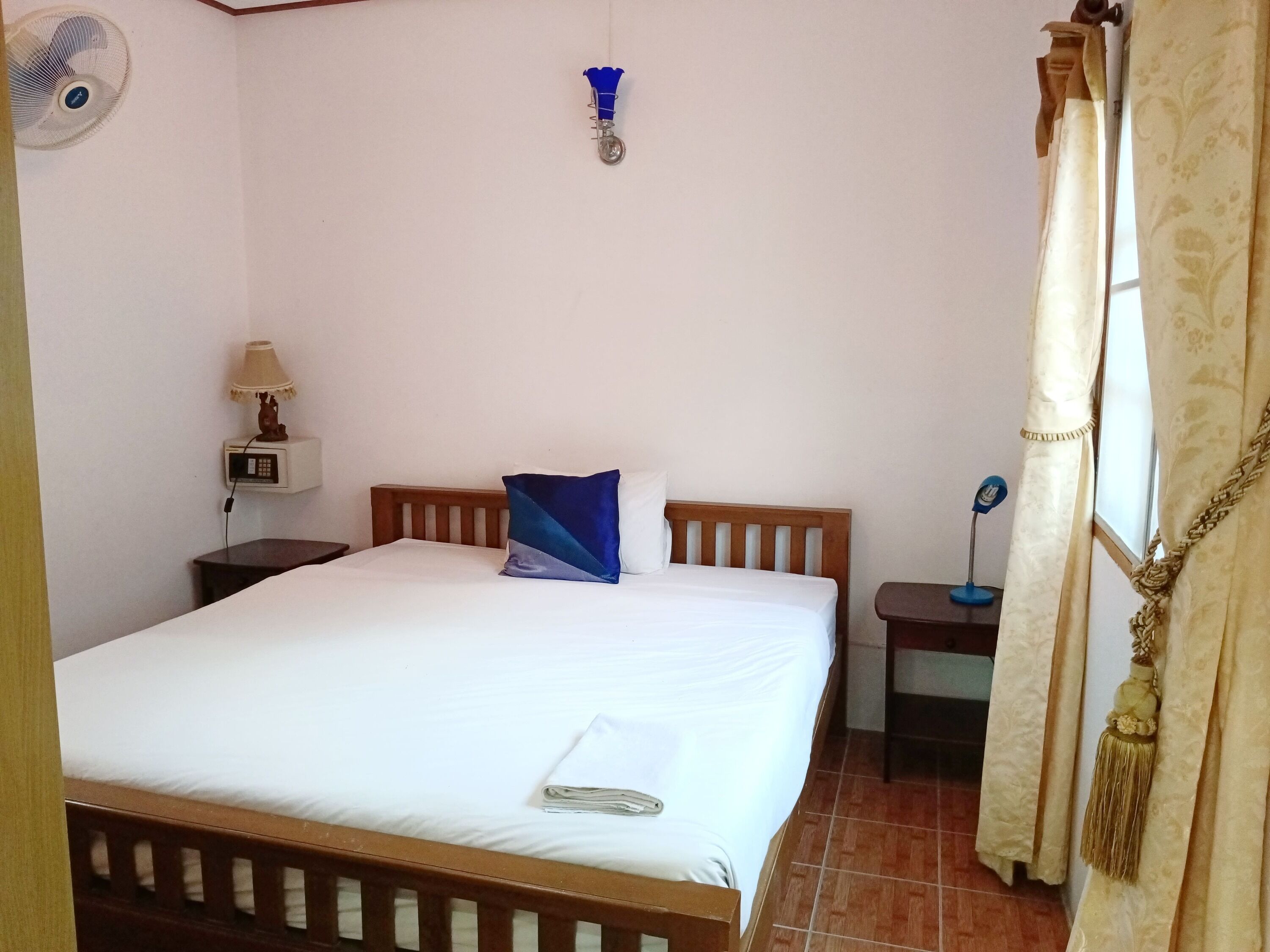 A bedroom with a wooden bed, white bedding, a blue pillow, a bedside table with a lamp, and a wall-mounted fan.