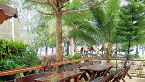An outdoor seating area with wooden benches and tables, surrounded by lush greenery and palm trees. - Sunrise Resort (Bang Saphan)