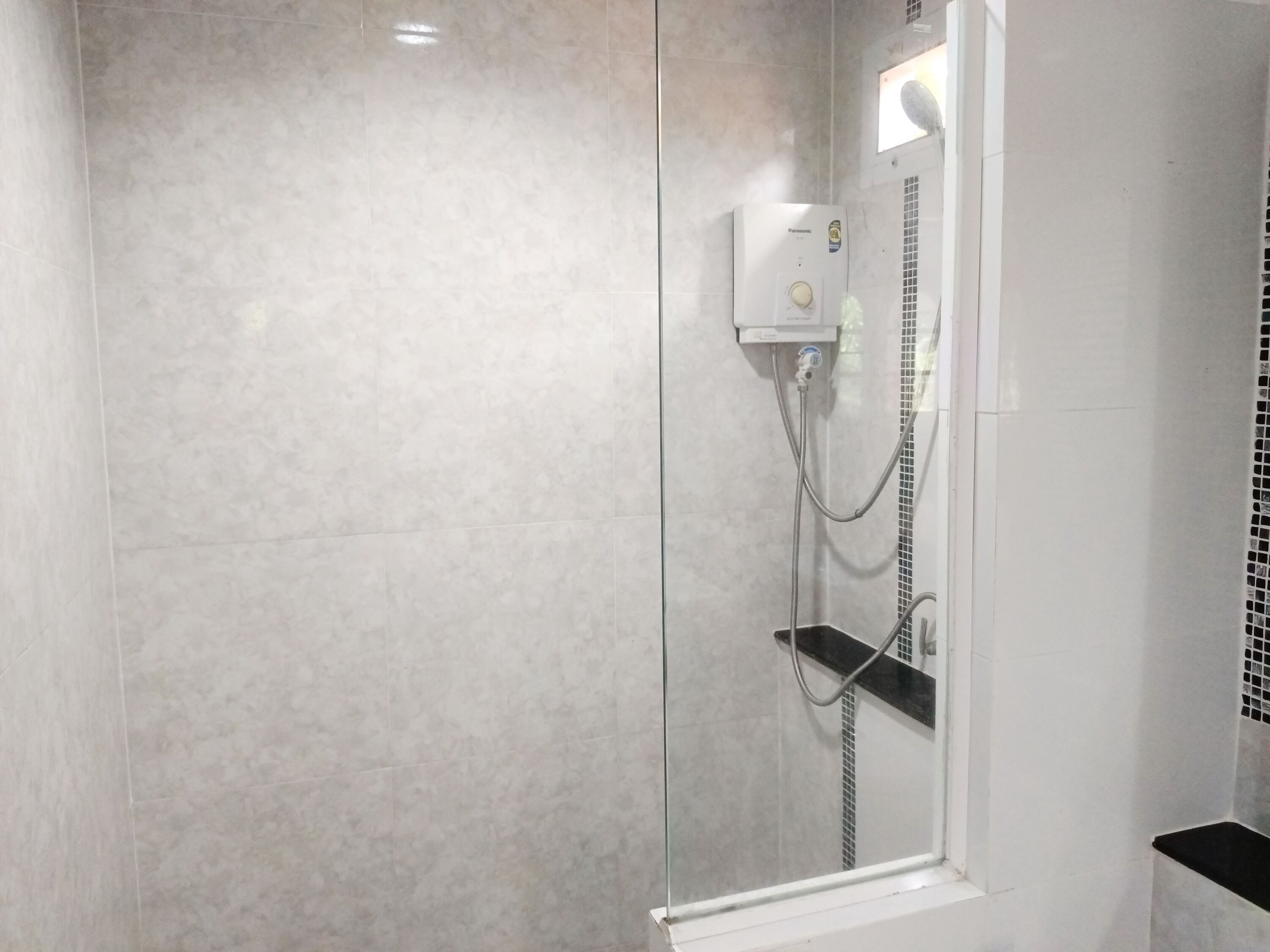 Un baño moderno con ducha separada por mampara de vidrio, pared de azulejos blancos y un calentador de agua Panasonic visible.