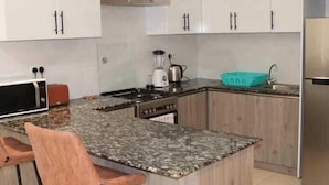 A kitchen with white cabinets, a granite countertop, a stove, a sink, and two bar stools. - Lux Suites Silverstone  Apartments (Nairobi)