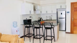A modern kitchen with white cabinets, a stainless steel refrigerator, and a water dispenser. There are three black bar stools at the island. - Lux Suites Silverstone  Apartments (Nairobi)