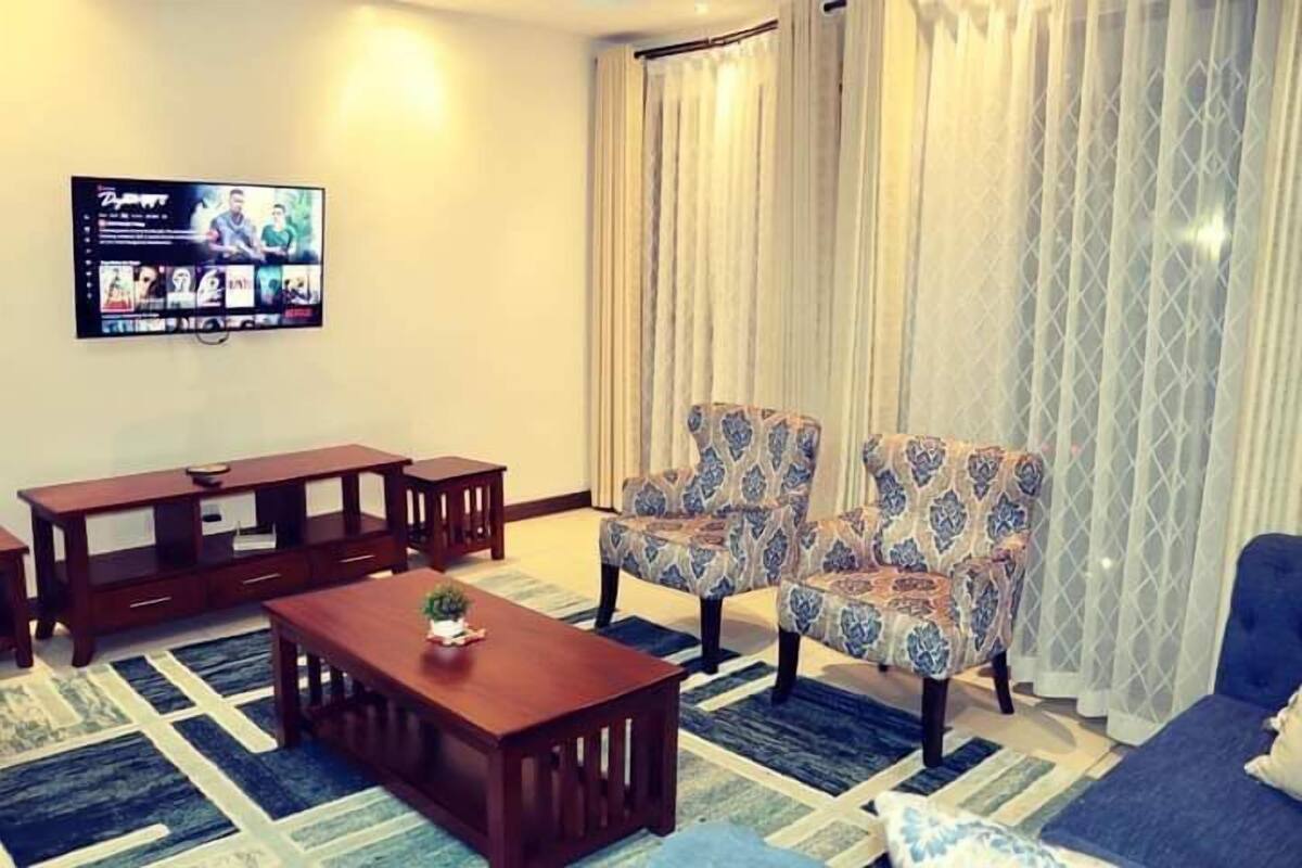 A living room with a TV mounted on the wall, a wooden coffee table, two patterned armchairs, a blue sofa, and a patterned rug.
